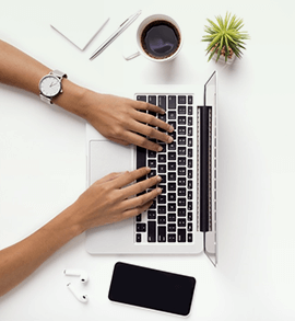 Top view of hands typing on a laptop keyboard, surrounded by a coffee cup, smartphone, notepad, pen, wireless earbuds, and a small potted plant on a white desk.